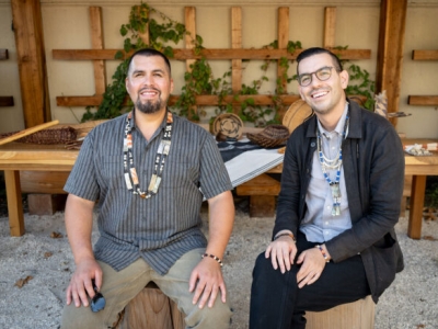 Two people sit on chairs made from tree trunks and smile at the camera. Behind them is a long wooden table that holds basketry and serving utensils.