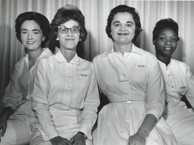 A black and white photo of three white woman and one Black woman, all with 1960s hair and wearing white uniforms.