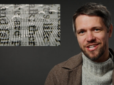 A smiling man on a gray background with an inset image of unreadable text