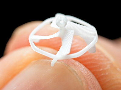 A photo of a small, white, propeller-shaped object that is held between the tips of two fingers. The object is smaller than the width of a fingernail.