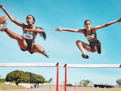 Two young women athletes are pictured in midair as they leap over hurdles. One is slightly ahead and both have expressions of intense concentration.