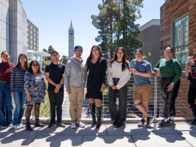 Nine students stand outside on the UC Berkeley campus, with the campanile visible in the background.