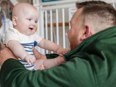 a man in a green hoodie holding a smiling baby with both arms