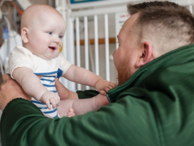 a man in a green hoodie holding a smiling baby with both arms