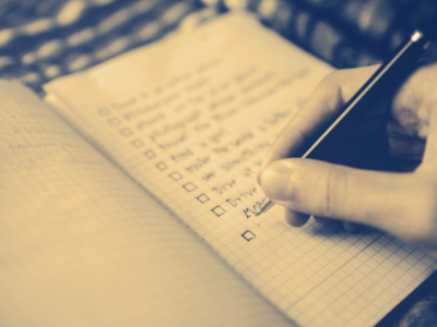 A person's hand writing a list of items on a piece of paper next to boxes that will be checked off.