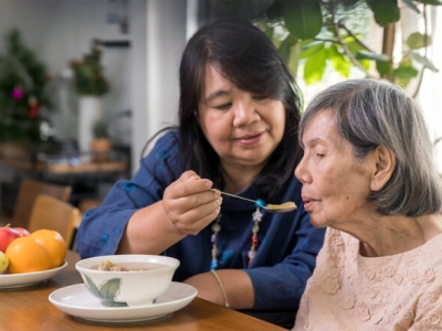 a female caregiver, apparently of middle age, feeds an elderly woman with a spoon