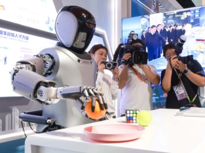 A silver- and black-colored humanoid robot grasps an orange with a mechanical hand. Three people stand behind the robot taking photos.