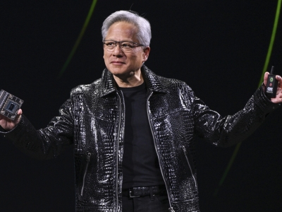 An Asian man with glasses and short gray hair wears a black shiny reptile leather jacket. His arms are outstretched and he holds a piece of electronic equipment in each hand.