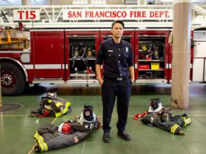 A photo of Maiko Bristow, a women firefighter with the San Francisco Fire Department. Maiko is wearing a black uniform and her turn out gear sits on the ground around her. Behind her is a fire truck with the words "San Francisco Fire"