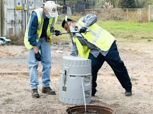 Two men in yellow safety vests stand outside on either side of an open sewer drain. Between them sits a wastewater autosampler, a large grey plastic cylinder about the size of a trashcan.