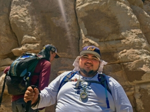 Morino Baca smiles at the camera wearing a cal hat standing on front of a rock cliff