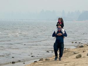 A man carries a small child along the beach of Lake Tahoe. They are both wearing face masks, and the air is cloudy with wildfire smoke.