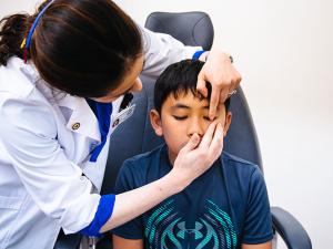 A photo shows a child sitting in an exam chair. An optometrist stands over the child, examining their eye.