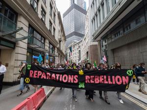 A photo shows protestors marching down a city street holding a large sign that reads "Reparations, Rebellion," in large, pink on black letters