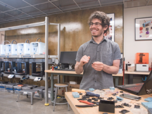 A photo of a student standing at at table, smiling, with a bank of 3D printers in the background.