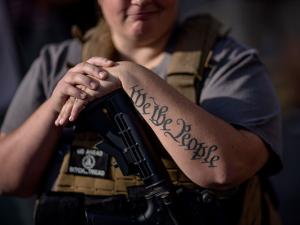 A smiling person with a large rifle and "We the People" tattooed on her arm