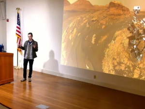 Sergey Levine speaks from a stage with a large screen projecting the Mars rover in the background