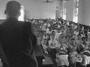 Black and white image of Martin Luther King Jr. with his back to the camera, and standing at a church pulpit speaking to a congregation of churchgoers sitting on church pews.
