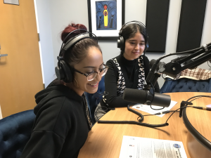 Two students smile as they speak into a microphone while sitting at a table. They are also wearing headphones and speaking as part of a podcast.