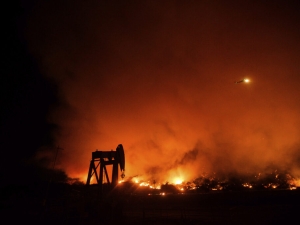orange flames from a wildfire in California burning at night in the distance, with the silhouette of an oil and gas pumpjack in the foreground.