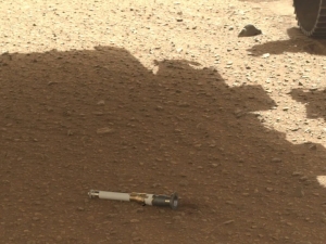 a small white and black tube sits on pebbly red ground in the shade of the Insight lander on Mars