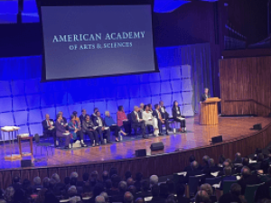 Multiple people, presumably scholars, sitting in chairs on the stage of an auditorium, overlooking an audience. One person is standing at a podium on the stage. A projected image behind the people on the stage reads "American Academy of Arts & Sciences." 