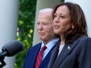 Vice President Kamala Harris speaks into a microphone outside of the White House, with a serious expression. President Joe Biden is to her side and slightly behind her, also looking serious.