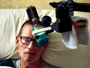 A robotic arm holding a brush raised over the head of a man lying in bed.