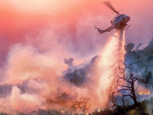A helicopter dropping water on a California wildfire in rugged terrain, backlit by a setting sun filtered through multiple layers of smoke.