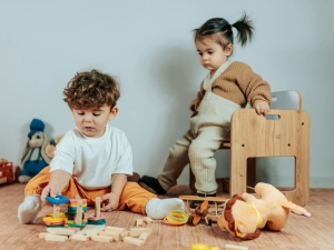 two toddlers in a room, with one looking over the shoulder of the other as they play with blocks. the child playing with the blocks has amassed several in front of him.