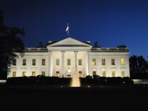 The White House in Washington D.C. at night, illuminated with lights and with a fountain in the foreground shooting water into the air.