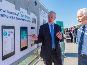 Richard Allen, in a suit, points to a large printout of smartphone screens showing how the MyShake app works.