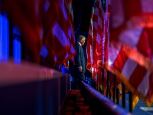 With a row of American flags on the left and right side of the frame, Donald Trump walks in the distance while supporters cheer
