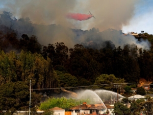 Smoke emerges from forested hills. In the foreground, two fire trucks aim streams of water at a vulnerable home. Overhead, an aircraft releases a plume of red flame retardant chemicals.