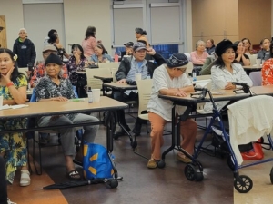 Various members of the Khmer community sit at rows of tables at a room. Many of them appear to be engaged in conversation. One wears a flower in her hair, while another is smiling serenely while wearing a fur hat.