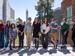 Nine students stand outside on the UC Berkeley campus, with the campanile visible in the background.