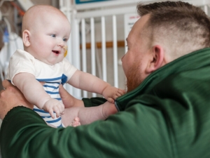 a man in a green hoodie holding a smiling baby with both arms