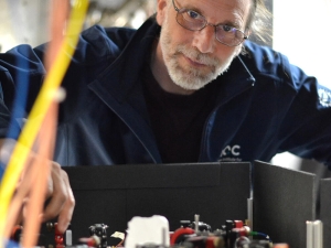 A man with glasses and a blue jacket tinkers with equipment while looking at the camera