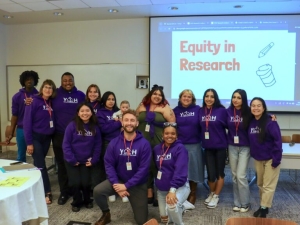 A group of 14 people who are involved in Coco Auerswald's project involving young people who experienced homelessness in the research process stand in front of a screen that reads 