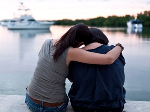 two women sitting at waters edge; one with are around the other's shoulders; both heads down